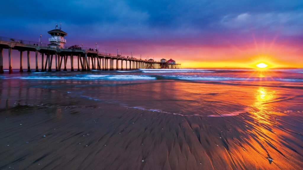A breathtaking sunset over Huntington Beach Pier in California, with vibrant colors reflecting on the ocean waves and sandy shore. The pier stretches out into the water, silhouetted against the colorful sky, creating a serene and picturesque coastal scene.