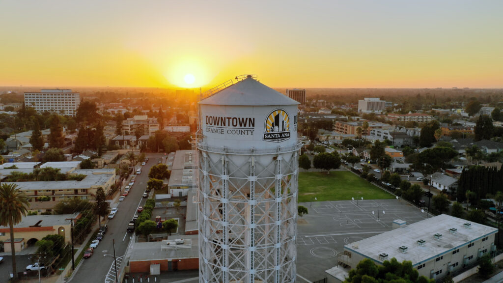 Aerial view of Downtown Santa Ana in Orange County at sunset, featuring a prominent water tower with "Downtown Orange County Santa Ana" written on it. The sun is setting behind the cityscape, casting a warm glow over the buildings, streets, and surrounding areas. The image captures the urban environment with a mix of residential and commercial buildings, along with a sports court visible in the foreground.