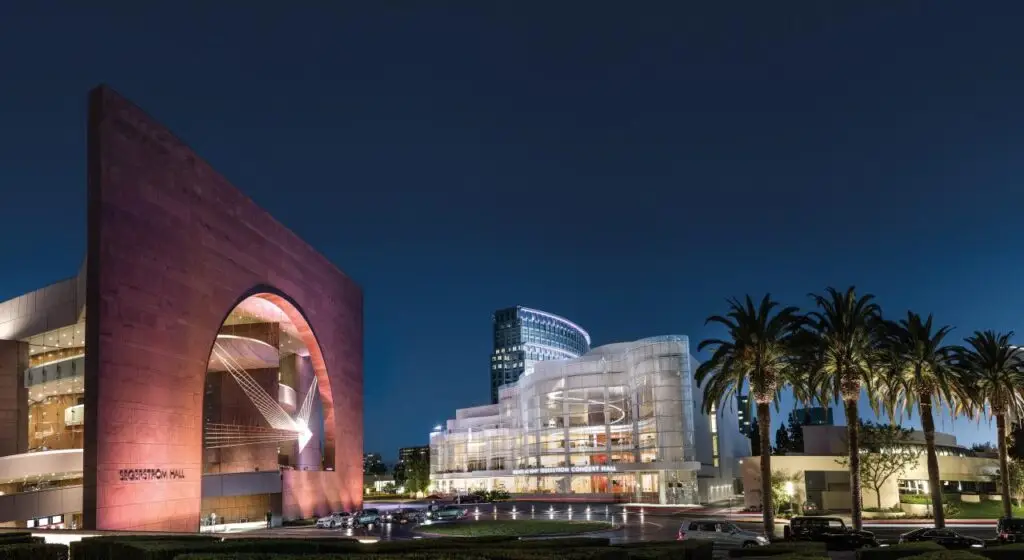 Night view of the Segerstrom Center for the Arts in Costa Mesa, California, featuring the illuminated red Segerstrom Hall and the modern glass facade of the Renee and Henry Segerstrom Concert Hall. Palm trees line the foreground, enhancing the elegant and sophisticated atmosphere.