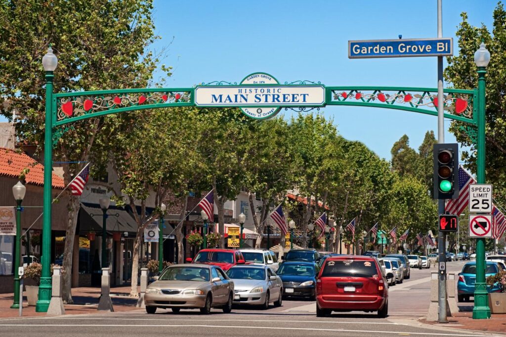 A vibrant scene of Historic Main Street in Garden Grove, California, featuring an archway sign with strawberries and "Historic Main Street" written on it. The street is lined with shops and adorned with American flags, creating a welcoming and patriotic atmosphere. Cars and pedestrians add to the bustling vibe of the area.