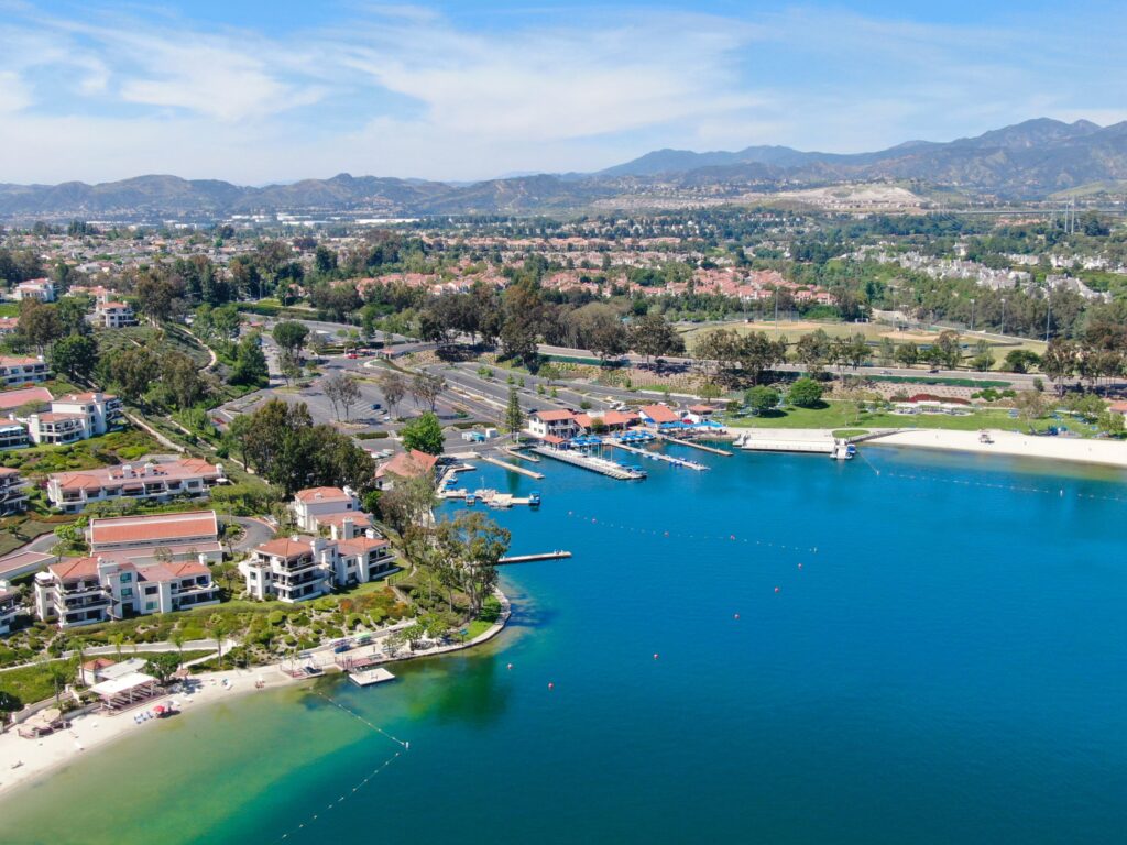 Aerial view of Lake Mission Viejo in Mission Viejo, California, featuring clear blue waters, sandy beaches, and Mediterranean-style residential areas. The surrounding lush green parks and distant rolling hills create a picturesque and serene environment under a clear blue sky.